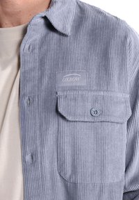 Camisa de pana gris claro con cuello, un bolsillo en el pecho y cierre de botones. Presenta una textura acanalada y un parche de OX-BOW.