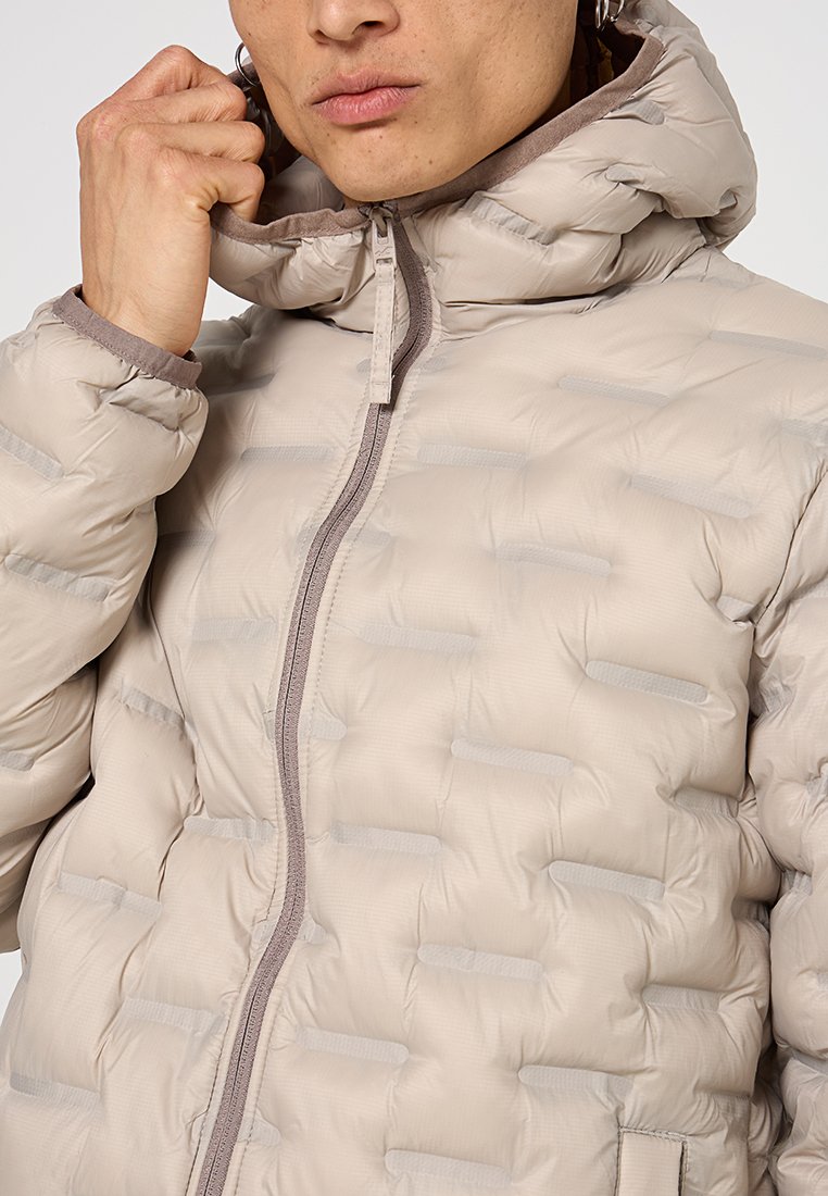 Osoba ubrana w beżową pikowaną kurtkę puchową z kapturem, trzymająca kołnierz blisko podbródka, częściowo widoczna twarz z neutralnym wyrazem.