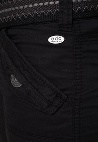 Les jeans en denim noir présentent une poche avant avec des détails de couture et une étiquette argentée. Le tissu a une texture lisse avec un léger éclat.