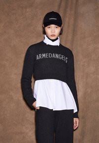 Suéter negro cortado con "ARMEDANGELS" en gris, superpuesto sobre una camisa blanca con botones. Combinado con pantalones oscuros y una gorra negra.