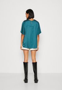 Teal satinbluse med korte ærmer, afslappet pasform, nøglehul detalje i ryggen, parret med shorts og knæhøje sorte støvler. Glat tekstur.