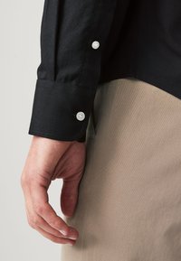 Close-up of a person's hand near the side of beige pants, wearing a black long-sleeve shirt with white buttons on the cuff and sleeve.