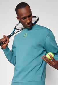 Sudadera azul verdosa con cuello redondo, puños acanalados y un pequeño logo amarillo. La persona sostiene una pelota de tenis y una raqueta en una pose relajada.