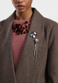Blazer a motivo houndstooth marrone sopra un maglione rosa, adornato con gemme multicolori e una spilla con frange di cristallo, caratterizzato da accenti metallici lucidi.