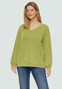 Vrouw in een lichtgroene V-hals sweatshirt en een blauwe jeans, staand tegen een effen lichte achtergrond met een lichte glimlach.