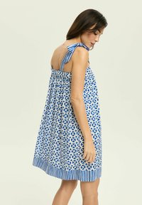 Niebieska i biała sukienka o geometrycznym wzorze, bezrękawna, z wiązanymi ramiączkami i paskowanym dołem. Materiał wydaje się lekki i zwiewny.
