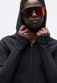 Άτομο που φοράει μαύρο φούτερ με κουκούλα και μεγάλα κόκκινα αθλητικά γυαλιά ηλίου με ανακλαστικούς φακούς, κρατώντας το φούτερ κοντά στον γιακά με τα δύο χέρια.