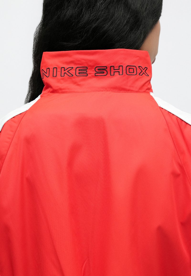 Rød jakke med høj krave, med sort "NIKE SHOX" tekst på ryggen og hvide accenter på skuldersømmene. Glat tekstur.