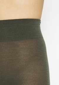 Calze opache di un verde scuro con un elastico a coste; lunghezza a metà coscia. Il tessuto è morbido e leggermente traslucido sotto l'elastico.
