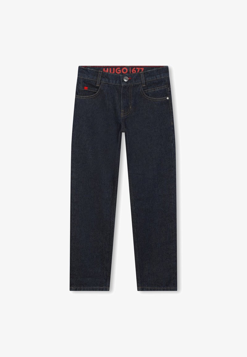 Un jean en denim bleu foncé présente une coupe droite, un design à cinq poches et des coutures de contraste. La ceinture affiche une étiquette de logo rouge.