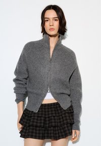 Junge Frau trägt einen grauen, gerippten Reißverschluss-Pullover und einen schwarzen, geschichteten karierter Rock, steht vor einem schlichten weißen Hintergrund.