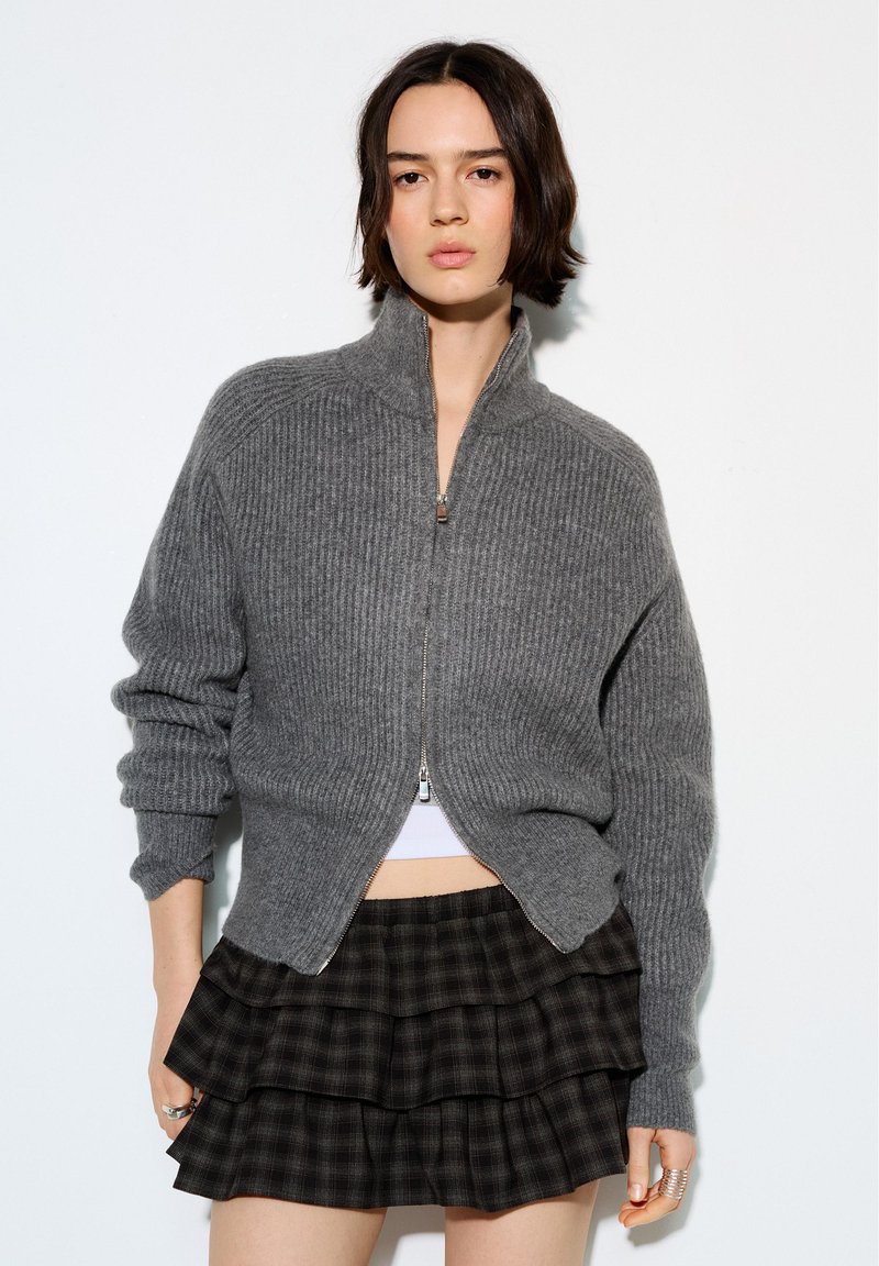 Junge Frau trägt einen grauen, gerippten Reißverschluss-Pullover und einen schwarzen, geschichteten karierter Rock, steht vor einem schlichten weißen Hintergrund.