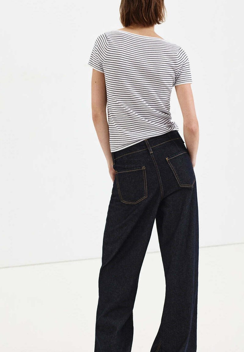 Femme portant un t-shirt rayé blanc et noir à manches courtes et un jean large foncé, debout, les mains dans les poches, vue de dos.