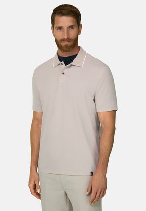 Homme barbu portant un polo beige à manches courtes et un pantalon clair, debout et faisant face à un fond uni.