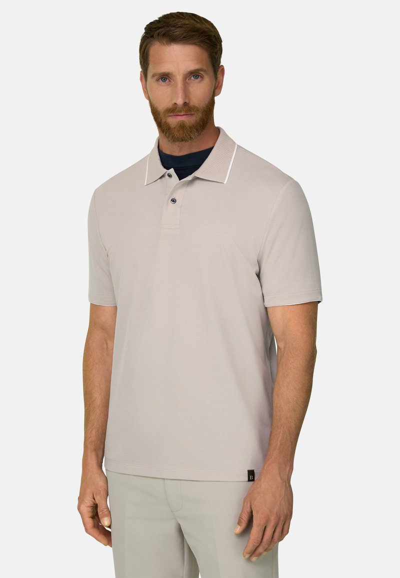 Homme barbu portant un polo beige à manches courtes et un pantalon clair, debout et faisant face à un fond uni.