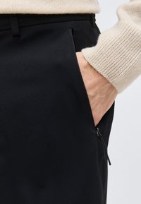 Χέρι στην τσέπη μαύρου παντελονιού με φερμουάρ, φορώντας μπεζ ριγωτό πουλόβερ με μανίκι.