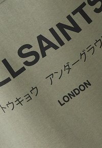 Maslinasto zeleni dukserica s velikim crnim natpisom, koji sadrži "ALLSAINTS" i "LONDON", uz manji višejezični tekst ispod. Meki, teksturirani materijal.