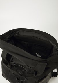 Črna tkaninska torba z debelimi naramnicami, prostornim notranjostjo in mrežnim žepom. V notranjosti je vidna majhna oznaka blagovne znamke. Poglej od zgoraj, z zadrgo.