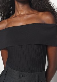 Bodycon nero a spalle scoperte con texture a costine e forma aderente, abbinato a pantaloni grigi a vita alta con dettaglio a bottone.