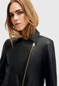 Zwarte leren jas met een hoge kraag, asymmetrische ritssluiting aan de voorkant met gouden hardware en zijzakken. Gladde textuur en nauwsluitend ontwerp.