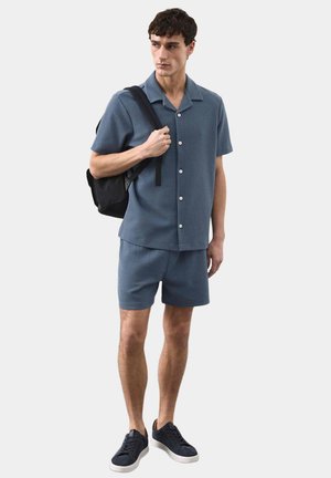 Młody mężczyzna w niebieskiej koszuli na krótki rękaw z guzikami i dopasowanych szortach, niosący czarny plecak przewieszony przez jedno ramię, stoi na jednolitym tle.