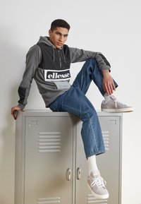 Felpa grigia e nera con logo, pantaloni grigi, sneakers grigie e calze bianche; posato su un armadietto metallico con una superficie testurizzata.