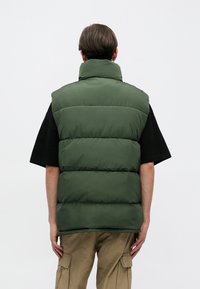 Roheline puhvvest kõrge kaelusega ja tikandiga disain. Kantud musta särgi peal, nähtav tagant, lühikeste juustega inimesel.