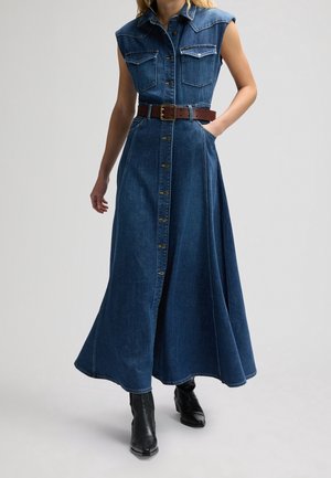 Kobieta w ciemnoniebieskiej, bezrękawnikowej sukience jeansowej z guzikami z przodu, kieszeniami na piersi, brązowym paskiem i czarnymi botkami na jasnym tle.