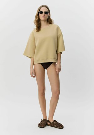 Vrouw met een beige oversized korte mouwsweatshirt, zwarte bikinibroekjes, bruine sandalen en zwarte ovale zonnebril tegen een effen lichte achtergrond.