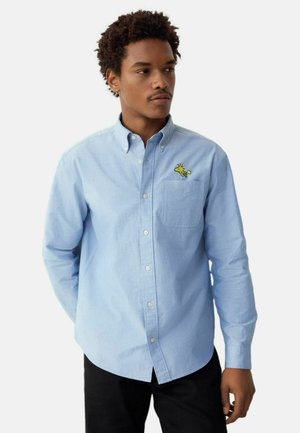 Light blue button-down shirt in cotton with long sleeves, a chest pocket, and a yellow embroidered character on the left side.