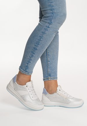 Persona que lleva vaqueros ajustados azul claro y zapatillas blancas bajas con detalles en azul claro, de pie con un pie ligeramente levantado.