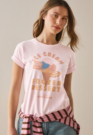 Kvinde iført en lyserød t-shirt med grafik af en isvaffel og tekst, kombineret med jeans og en lyserød og brunstribet sweater bundet om taljen.