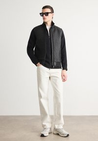 Μαύρο γιλέκο με ραβδωτή υφή πάνω από μαύρη ζακέτα με φερμουάρ, συνδυασμένο με άσπρο παντελόνι και γκρίζα sneakers, αξεσουάρε με μαύρα γυαλιά ηλίου.