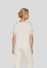 Helles beiges Hemd mit kurzen Ärmeln, das mit Spitzen Details an den Bündchen verziert ist. Weicher Stoff mit einer lockeren Passform, ergänzt durch cremefarbene Hosen.