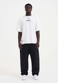 White oversized t-shirt featuring black graphic text, paired with loose black denim pants. White sneakers complete the outfit.
