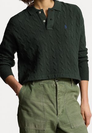 Pull en maille torsadée vert foncé avec col et boutons, porté avec un pantalon taille haute vert olive, mains dans les poches.