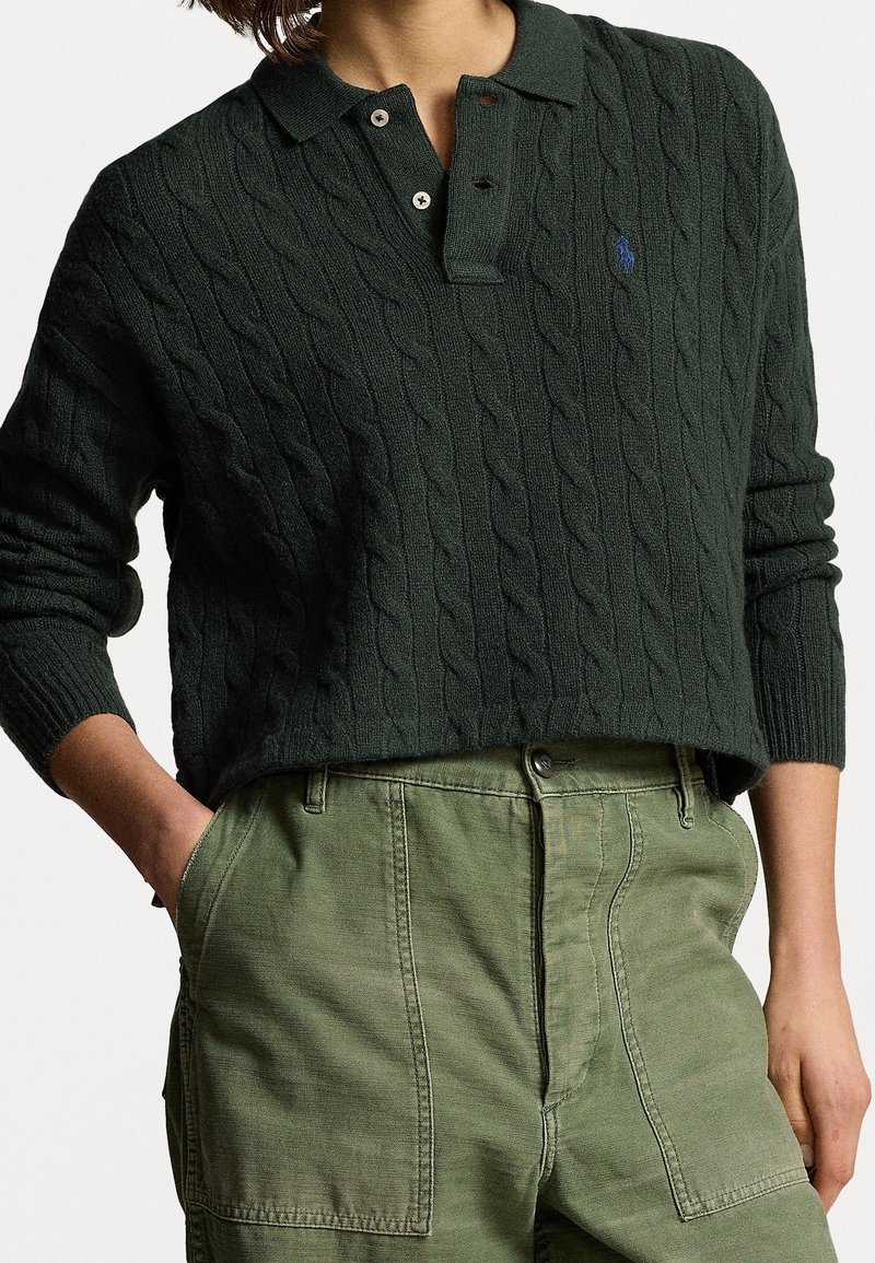 Pull en maille torsadée vert foncé avec col et boutons, porté avec un pantalon taille haute vert olive, mains dans les poches.