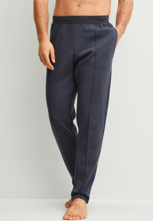 Man draagt donkergrijze joggingbroek, staat blootsvoets met één hand in de zak tegen een lichte achtergrond.