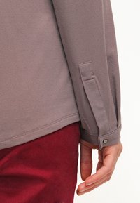 Langärmliges Hemd aus hellbraunem Stoff, mit einer schmalen Passform und einer geknöpften Manschette sowie einem Schlitzdetail an der Seite; getragen über roten Hosen.