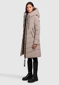 Vrouw in een lange beige gewatteerde jas met capuchon, zwarte leggings en hoge zwarte plateau laarzen, staand in profiel op een lichtgrijze achtergrond.