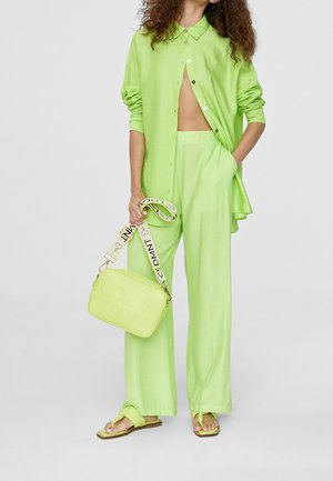 Limegrøn skjorte med knapper, matchende brede bukser, flip-flops og en rektangulær håndtaske med et mønstret реме, der står "LOLA CSDNT."