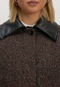 Bruine tweedjas met een zwarte leren kraag, sluiting met één knop en een gestructureerde stof. Zichtbare patroonvariaties verbeteren het ontwerp.