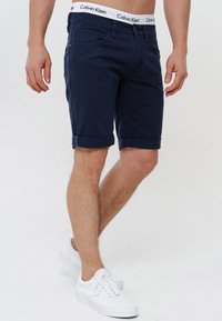 Muške tamnoplave traper šortse s preklopljenim rubovima, koji imaju prednje džepove i Calvin Klein brendiranu pojase. Uparene su s bijelim tenisicama.