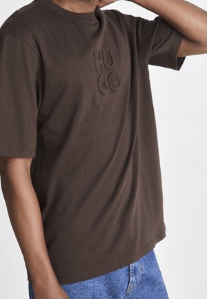 Personne portant un T-shirt marron de la marque Hugo associé à un jean denim bleu, debout devant un fond clair uni.