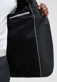 Schwarzes, maßgeschneidertes Sakko mit glattem schwarzem Futter, weißen Akzentnähten und einer Tasche mit weißem Streifendetail.