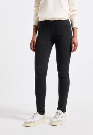 Leggings neri elasticizzati con una texture liscia, design a vita alta e senza chiusure visibili, abbinati a scarpe da ginnastica bianche e beige.