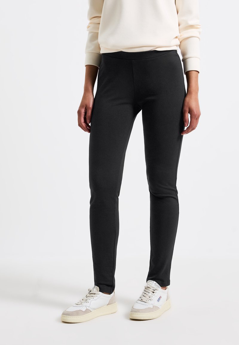 Svarta tajta leggings med en slät textur, hög midja och utan synliga knäppningar, i kombination med vita och beige sneakers.