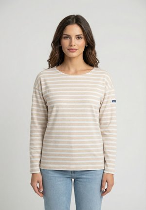 Mujer con cabello castaño ondulado, vestida con una camiseta de manga larga beige y blanca a rayas y vaqueros azul claro, de pie frente a un fondo liso.