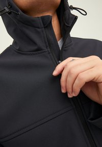 Chaqueta softshell negra con cuello alto, cierre de cremallera y capucha con cordón. Presenta una textura suave y detalles de costura visibles.