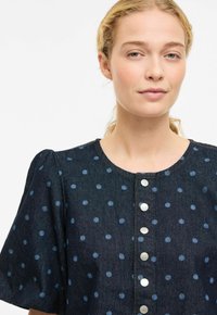 Chemise en denim foncé avec manches bouffantes, pois bleus et boutons-pression argentés sur le devant. Col rond simple et tissu texturé.